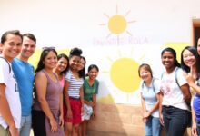 Um grupo diversificado de voluntários sorrindo e trabalhando juntos para pintar uma escola em uma comunidade rural, com um belo pôr do sol ao fundo.