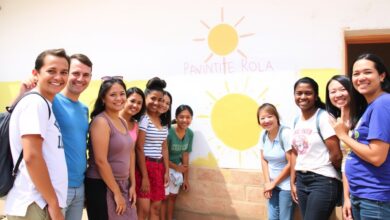 Um grupo diversificado de voluntários sorrindo e trabalhando juntos para pintar uma escola em uma comunidade rural, com um belo pôr do sol ao fundo.