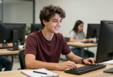 Jovem universitário(a) sorrindo enquanto trabalha em um escritório moderno como estagiário(a), com computador e anotações ao redor