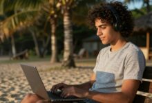 Jovem profissional com laptop trabalhando em praia exótica ao entardecer, representando liberdade de trabalhar viajando